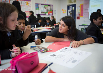 A female teacher crouches at the table with two students in an upper primary class in the UK. The students in the classroom are solving puzzles in pairs and have material on their tables to make something with paper and glue.