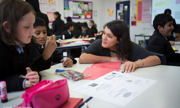 A female teacher crouches at the table with two students in an upper primary class in the UK. The students in the classroom are solving puzzles in pairs and have material on their tables to make something with paper and glue.