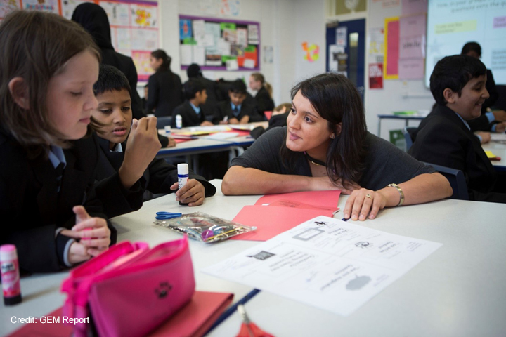 A female teacher crouches at the table with two students in an upper primary class in the UK. The students in the classroom are solving puzzles in pairs and have material on their tables to make something with paper and glue.