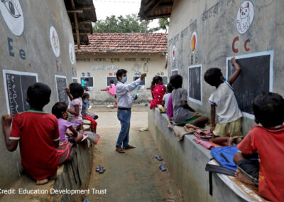 Male teacher coordinates children learning to write letters of the alphabet on the outside school walls during COVID-19, India.