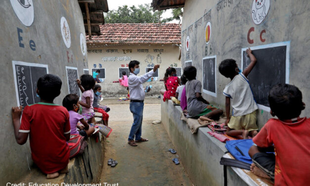 Male teacher coordinates children learning to write letters of the alphabet on the outside school walls during COVID-19, India.