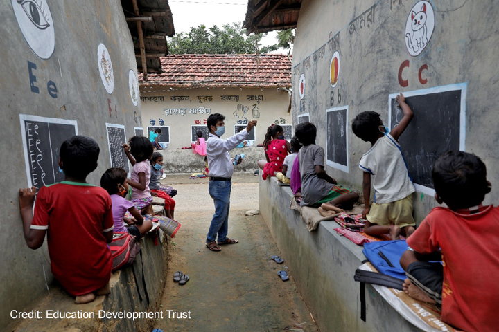 Male teacher coordinates children learning to write letters of the alphabet on the outside school walls during COVID-19, India.