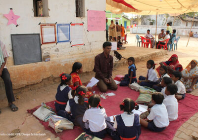 Teacher and community members lead a discussion with a class sitting on a blanket outside in the shade.