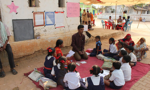 Teacher and community members lead a discussion with a class sitting on a blanket outside in the shade.