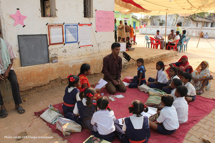 Teacher and community members lead a discussion with a class sitting on a blanket outside in the shade.