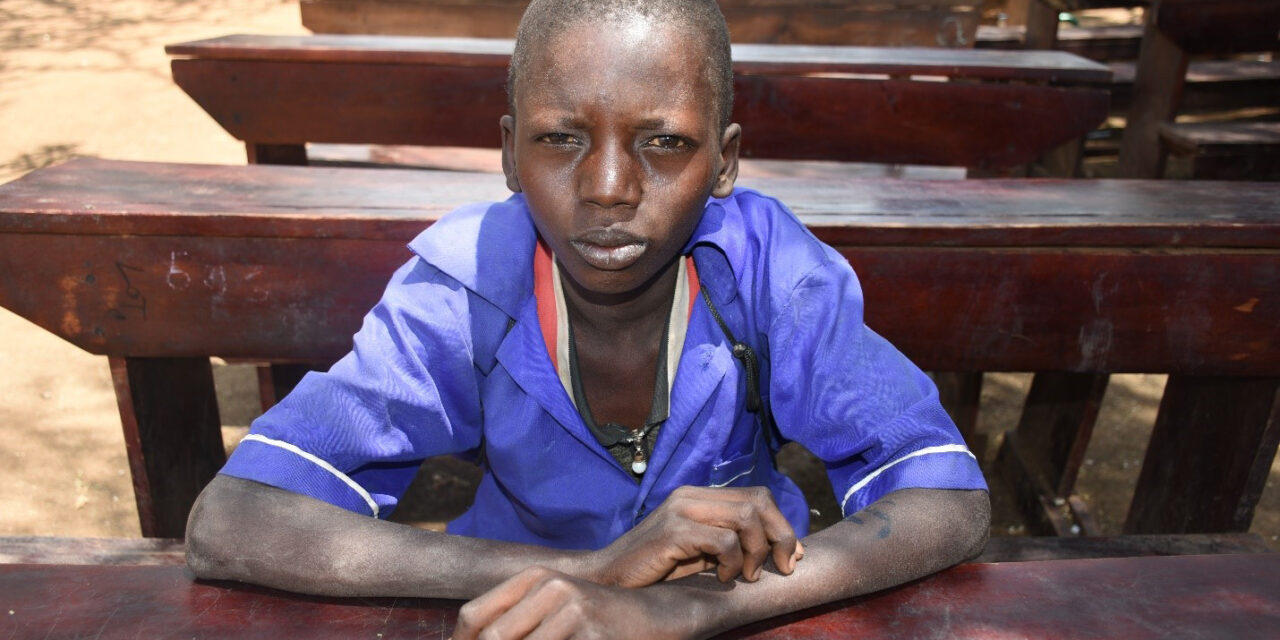 Image of Sagal, a boy sitting on a desk in the school yard. Sagal lives with his mother and three siblings in North East Uganda. The region is experiencing severe drought and many families are struggling to feed their children. Save the Children is providing school meals to help children stay in school and continue to learn.