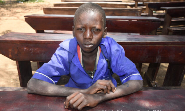 Image of Sagal, a boy sitting on a desk in the school yard. Sagal lives with his mother and three siblings in North East Uganda. The region is experiencing severe drought and many families are struggling to feed their children. Save the Children is providing school meals to help children stay in school and continue to learn.
