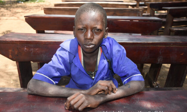 Image of Sagal, a boy sitting on a desk in the school yard. Sagal lives with his mother and three siblings in North East Uganda. The region is experiencing severe drought and many families are struggling to feed their children. Save the Children is providing school meals to help children stay in school and continue to learn.