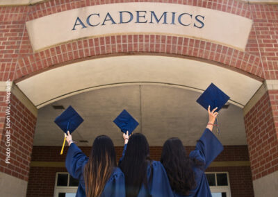 Three young women dressed in university graduation gowns hold up their graduation mortar boards looking to a sign saying ‘Academics’.