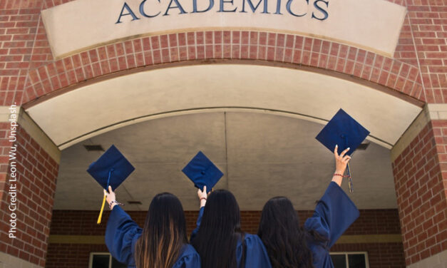 Three young women dressed in university graduation gowns hold up their graduation mortar boards looking to a sign saying ‘Academics’.
