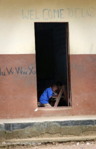  A young boy sits at his desk in a classroom in rural Sierra Leone and is visible through the open door