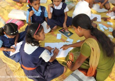 Children sit in small group circles on the floor assisted by teachers. This group shows a female teacher sitting on the ground helping a group of four girls, India.