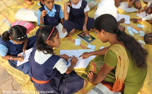 Children sit in small group circles on the floor assisted by teachers. This group shows a female teacher sitting on the ground helping a group of four girls, India.