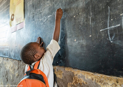 A small boy from Tanzania, reaching up to write on blackboard - big stretch - with orange rucksack