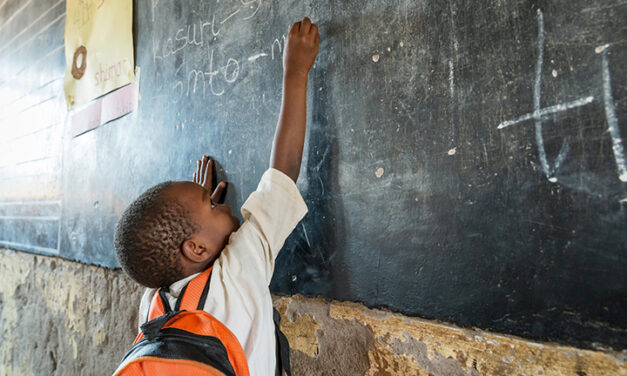 A small boy from Tanzania, reaching up to write on blackboard - big stretch - with orange rucksack