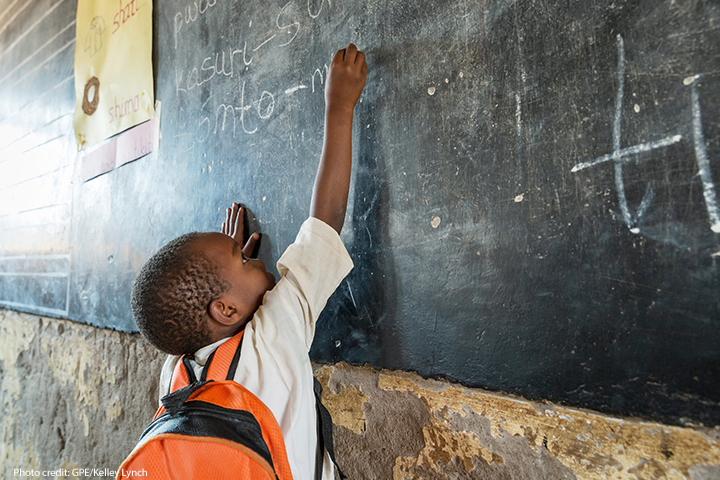 A small boy from Tanzania, reaching up to write on blackboard - big stretch - with orange rucksack