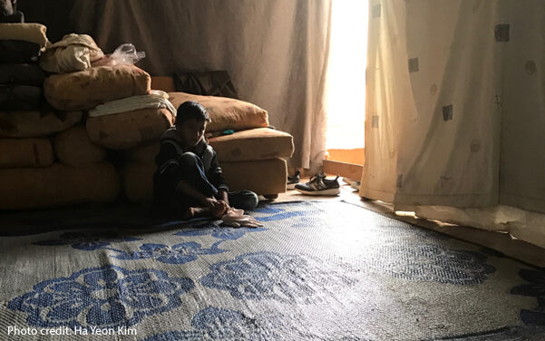 A Syrian refugee boy seated in a dimly-lit tent waiting his turn for social and emotional assessment