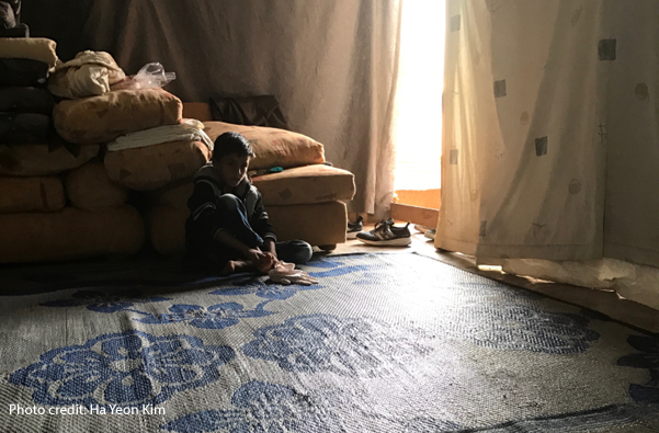 A Syrian refugee boy seated in a dimly-lit tent waiting his turn for social and emotional assessment