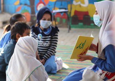 A small group of children in Pakistan sit on the floor discussing a book during Covid times, all wearing masks.