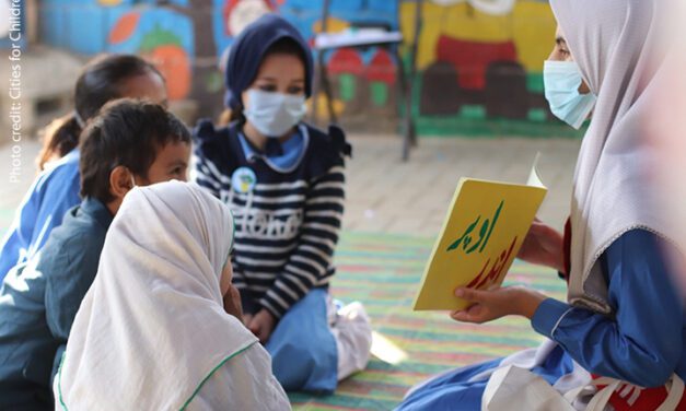 A small group of children in Pakistan sit on the floor discussing a book during Covid times, all wearing masks.