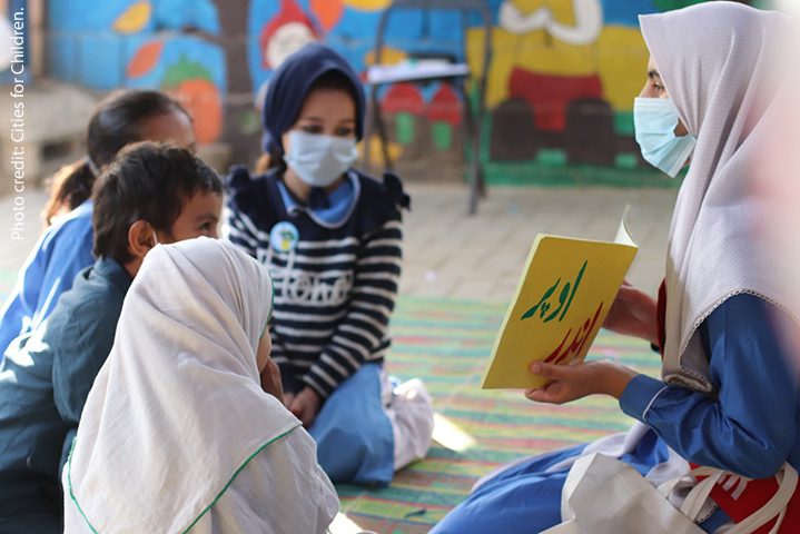 A small group of children in Pakistan sit on the floor discussing a book during Covid times, all wearing masks.