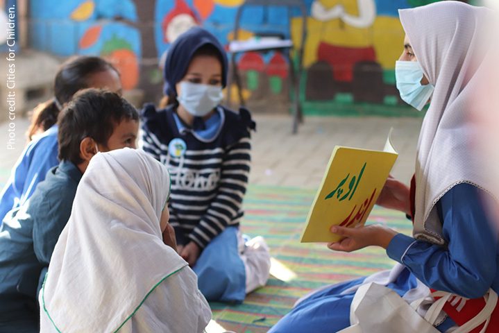 A small group of children in Pakistan sit on the floor discussing a book during Covid times, all wearing masks.