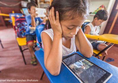 A girls sits in class watching a video on a tablet. She has her elbows on the desk with her hands over her ears