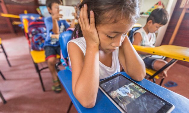 A girls sits in class watching a video on a tablet. She has her elbows on the desk with her hands over her ears