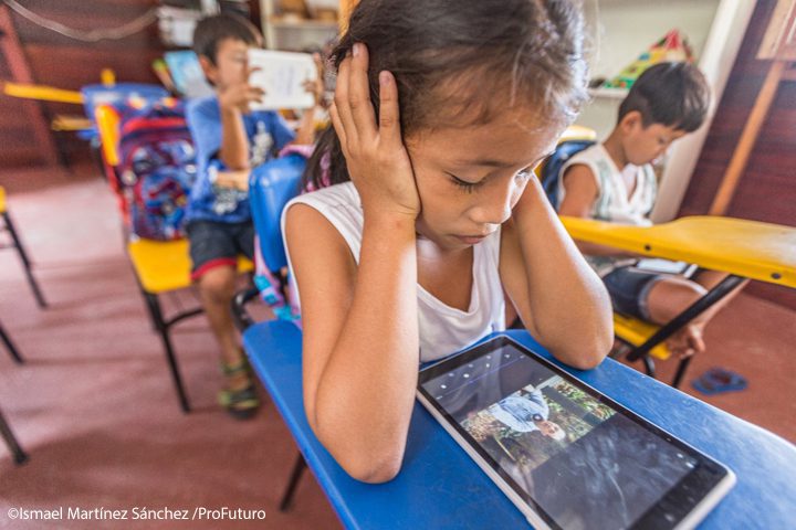 A girls sits in class watching a video on a tablet. She has her elbows on the desk with her hands over her ears