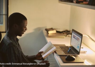 A male teacher sits at a desk studying with a book and laptop in the evening light