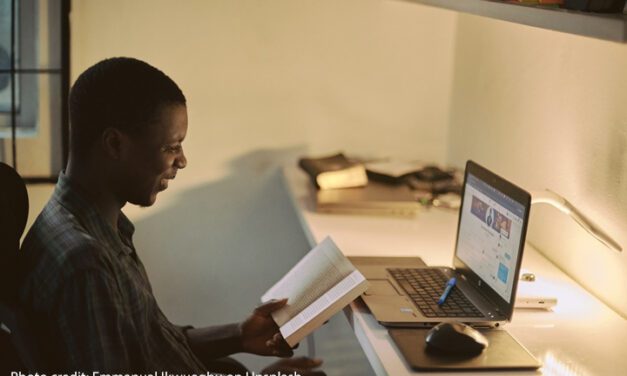 A male teacher sits at a desk studying with a book and laptop in the evening light