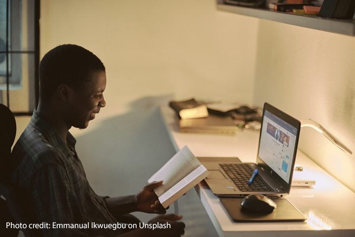 A male teacher sits at a desk studying with a book and laptop in the evening light