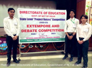 Project Voices team standing in front of a competition banner.  Banner reads: Directorate of Education Govt. of NCT of Delhi.  State level "Project voices" competition (2022-23) Extempore and debates competition 17 August 2023 Spoken English cell school branch 