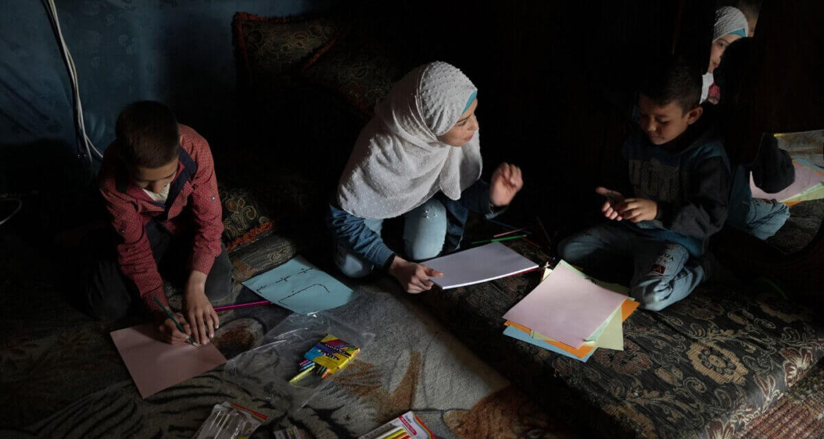 Children and a caregiver implement the remote early learning program at home on a carpet on the floor of a dark room with poor evening light.