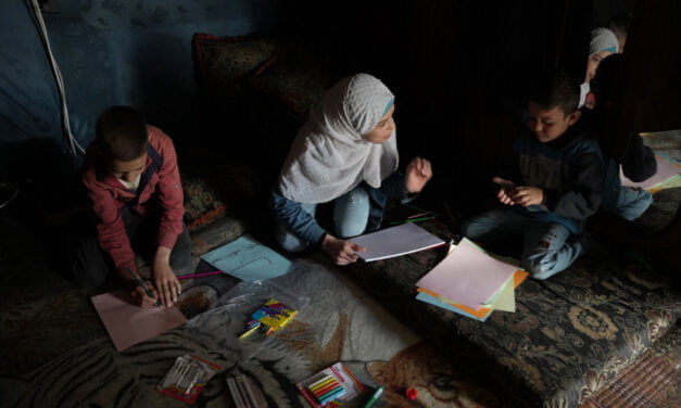 Children and a caregiver implement the remote early learning program at home on a carpet on the floor of a dark room with poor evening light.