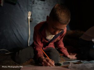 A boy is drawing as part of the the remote early learning program at home on a carpet on the floor of a dark room with poor evening light.