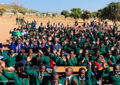 Children waving and wearing green uniform at the new Mushili Hillside secondary school celebrate outside with the Zambia Minister of Education and the UK Foreign Secretary.