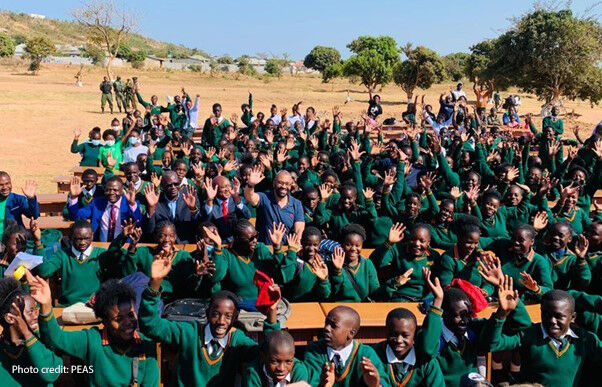 Children waving and wearing green uniform at the new Mushili Hillside secondary school celebrate outside with the Zambia Minister of Education and the UK Foreign Secretary.