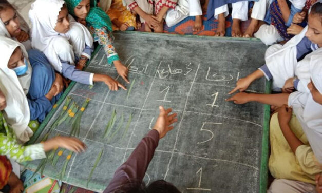 A para-teacher in Shikarpur using fallen leaves to teach his class the concept of ones and tens on a chalk board laid flat