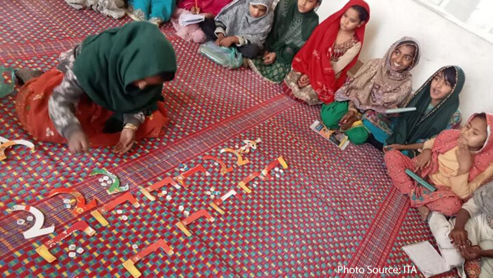 A para-teacher with children on a bright carpet   in Ghotki using used paper to show students the concept of nuqta (dot) in Sindhi Language.  