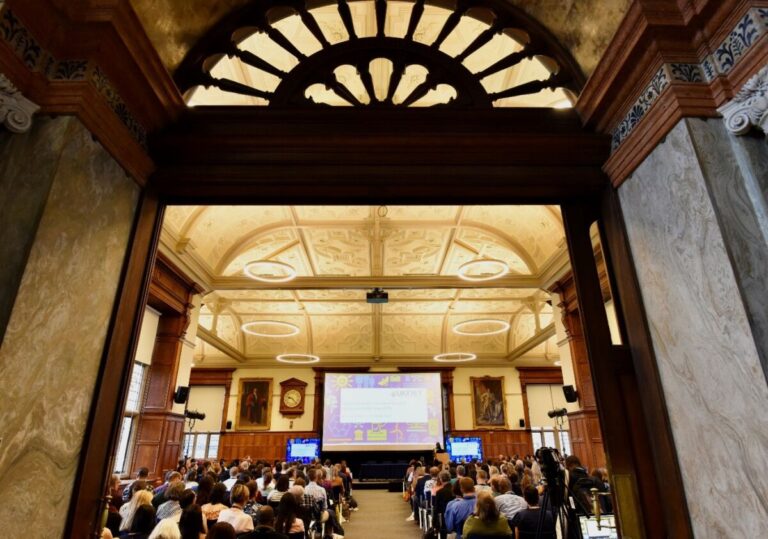 A view from the back of the room towards the screen in the UKFIET 2023 closing plenary. Picture also shows the arched window above the doorway and the ornate ceiling