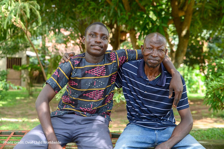 Young Ugandan man sitting with his arm around the shoulder of his father.  "Oyo, a 19-year-old deaf boy, sits with his father Oyo. Since Oyo has learned some sign language they have been able to have conversations. "