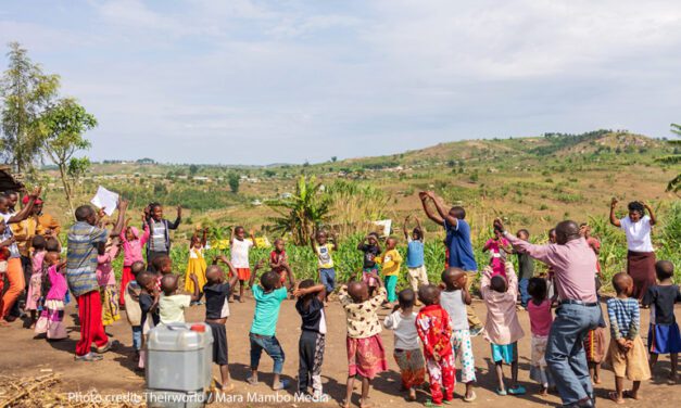 Colourfully dressed children play with their teachers in a circle, rural Uganda.