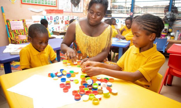 Sitting at a table with two children in yellow shirts, Kindergarten teacher, Madam Rebecca Antwi, engaging students in play-based learning at Obosomase Methodist School in Ghana's Eastern Region.