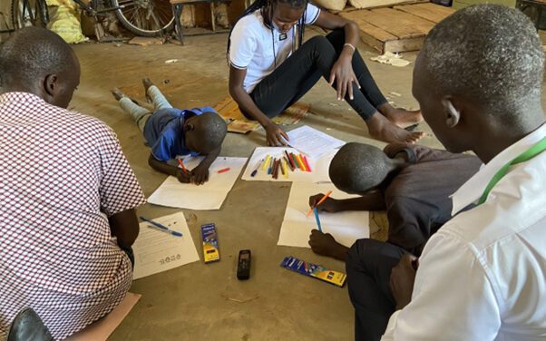 Children with disabilities engage in participatory research facilitated by peer researchers in Kenya. A group of adults and children working on the floor with coloured pens and paper