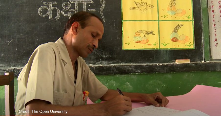 A male teacher writes in a classroom. Taken through the UK-Aid-funded TESS-India project, and a still from a video about TESS-India.