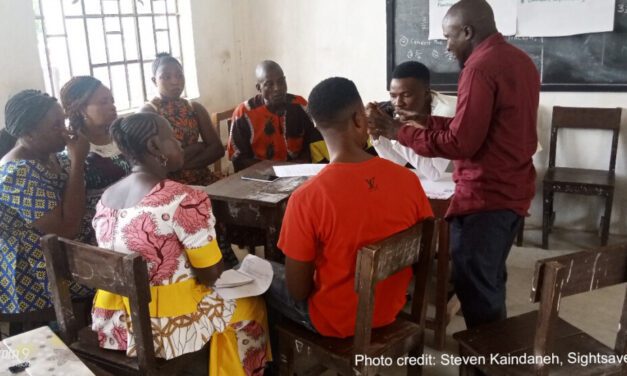 A group of teachers sit around a table in a classroom, engaging in a training workshop, Sierra Leone.