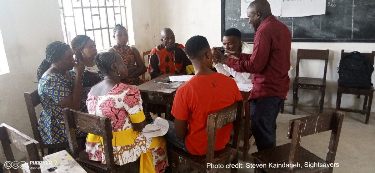 A group of teachers sit around a table in a classroom, engaging in a training workshop, Sierra Leone.