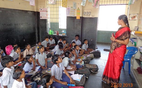 A female teacher stands at the front of the class with her students sitting on the floor in Chennai, India.