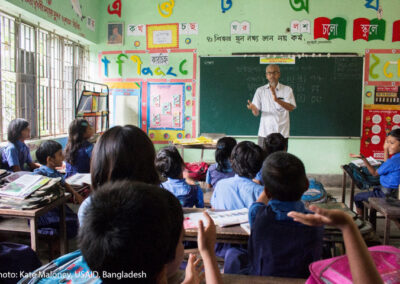 A male teacher stands at the front of a packed class of smiling students, Bangladesh.